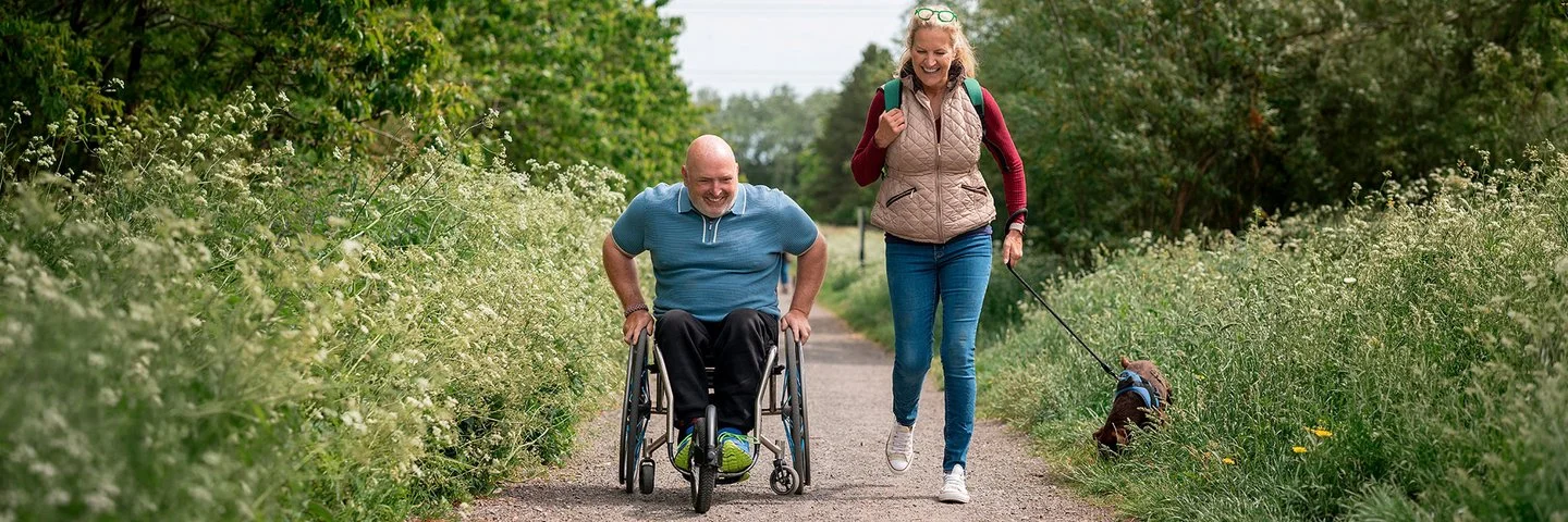 Ein Mann im Rollstuhl und eine Frau mit Hund sind gemeinsam auf einem Naturweg unterwegs.