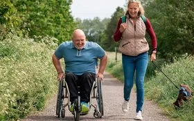 Ein Mann im Rollstuhl und eine Frau mit Hund sind gemeinsam auf einem Naturweg unterwegs. 