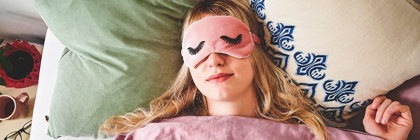 Eine junge Frau liegt im Bett und trägt eine Schlafmaske.