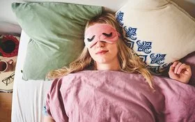 Eine junge Frau liegt im Bett und trägt eine Schlafmaske.
