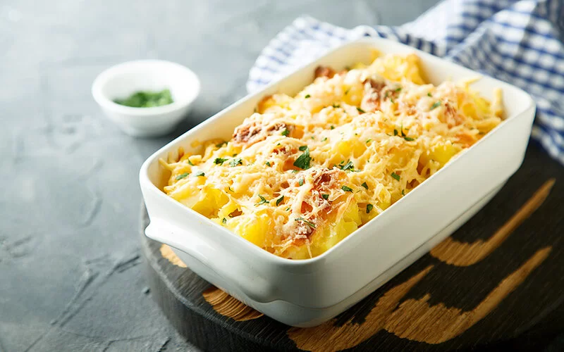 Auf einem Tisch steht eine Auflaufform mit überbackenen Kartoffeln. Der geriebene Käse darauf ist knusprig braun gebacken. Darüber sind frische Kräuter gestreut.