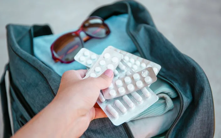 Eine Hand hält mehrere Medikamentenblister. Im Hintergrund ist eine gepackte offene Reisetasche abgebildet.