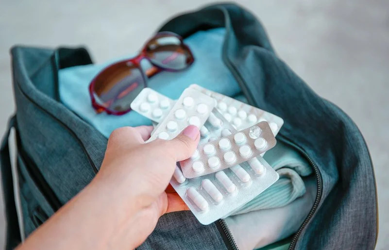 Eine Hand hält mehrere Medikamentenblister. Im Hintergrund ist eine gepackte offene Reisetasche abgebildet.