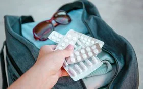Eine Hand hält mehrere Medikamentenblister. Im Hintergrund ist eine gepackte offene Reisetasche abgebildet.
