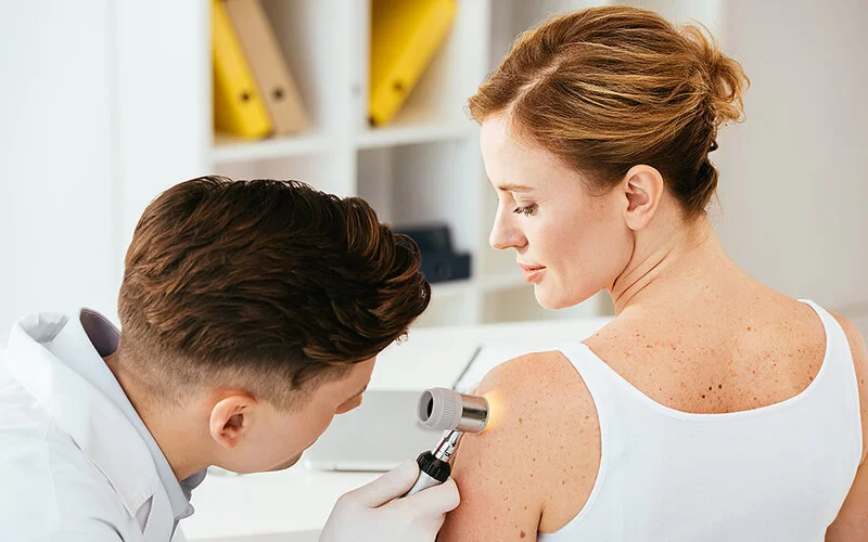 Ein junger Arzt untersucht mit einem Dermatoskop die Pigmentmale am Oberarm einer Patientin.