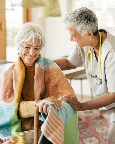 Eine ältere Frau sitzt mit einer Decke über den Schultern auf einem Stuhl. Ihre Hände stützen sich auf einen Gehstock. Eine Pflegerin beugt sich von der Seite zu ihr herunter. Mit der rechten Hand fasst sie der Frau auf die Schulter und mit der linken auf deren linken Arm.