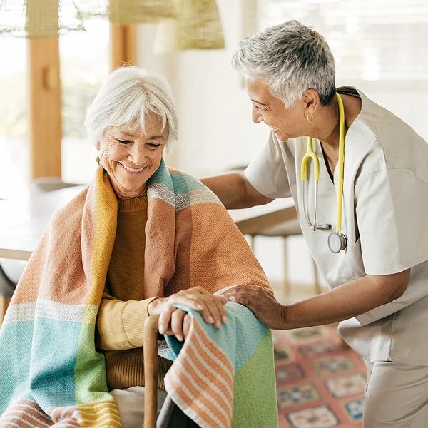 Eine ältere Frau sitzt mit einer Decke über den Schultern auf einem Stuhl. Ihre Hände stützen sich auf einen Gehstock. Eine Pflegerin beugt sich von der Seite zu ihr herunter. Mit der rechten Hand fasst sie der Frau auf die Schulter und mit der linken auf deren linken Arm.