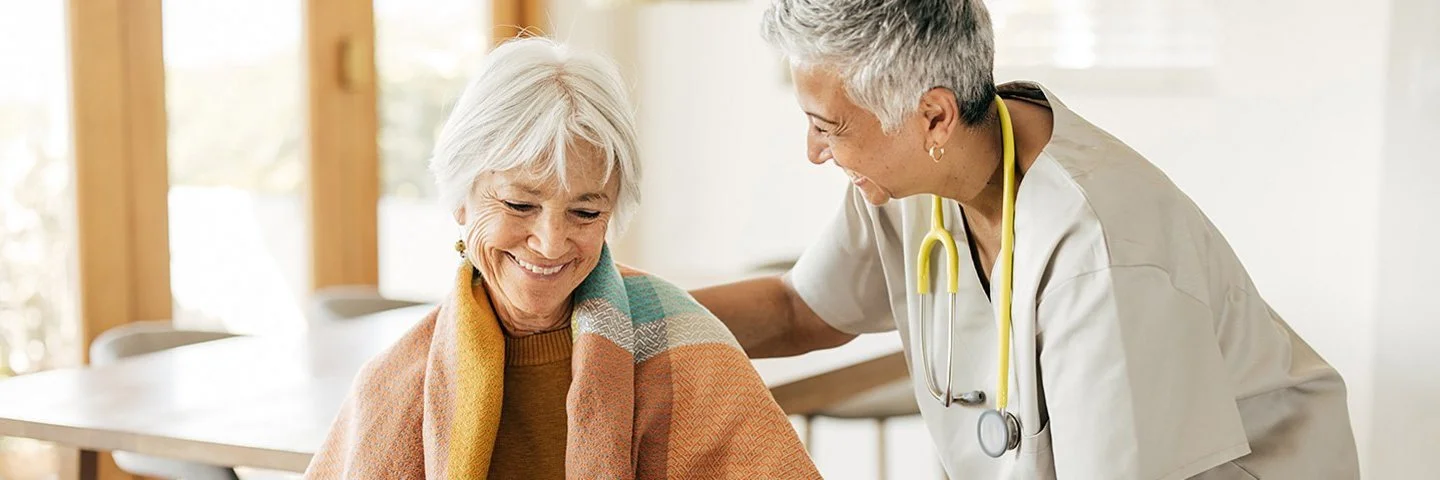 Eine ältere Frau sitzt mit einer Decke über den Schultern auf einem Stuhl. Ihre Hände stützen sich auf einen Gehstock. Eine Pflegerin beugt sich von der Seite zu ihr herunter. Mit der rechten Hand fasst sie der Frau auf die Schulter und mit der linken auf deren linken Arm.