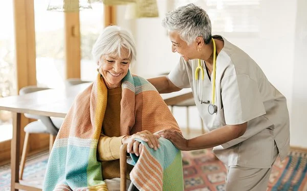 Eine ältere Frau sitzt mit einer Decke über den Schultern auf einem Stuhl. Ihre Hände stützen sich auf einen Gehstock. Eine Pflegerin beugt sich von der Seite zu ihr herunter. Mit der rechten Hand fasst sie der Frau auf die Schulter und mit der linken auf deren linken Arm.