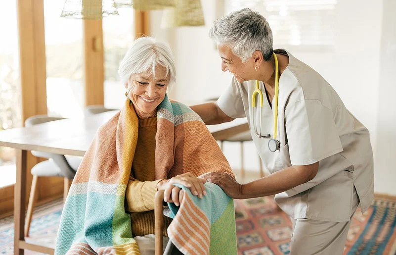 Eine ältere Frau sitzt mit einer Decke über den Schultern auf einem Stuhl. Ihre Hände stützen sich auf einen Gehstock. Eine Pflegerin beugt sich von der Seite zu ihr herunter. Mit der rechten Hand fasst sie der Frau auf die Schulter und mit der linken auf deren linken Arm.