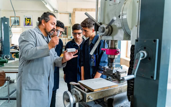 Ein Berufschullehrer erklärt in einer Feinmechanik-Werkstatt Auszubildenden ein Werkstück.