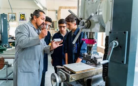 Ein Berufschullehrer erklärt in einer Feinmechanik-Werkstatt Auszubildenden ein Werkstück.