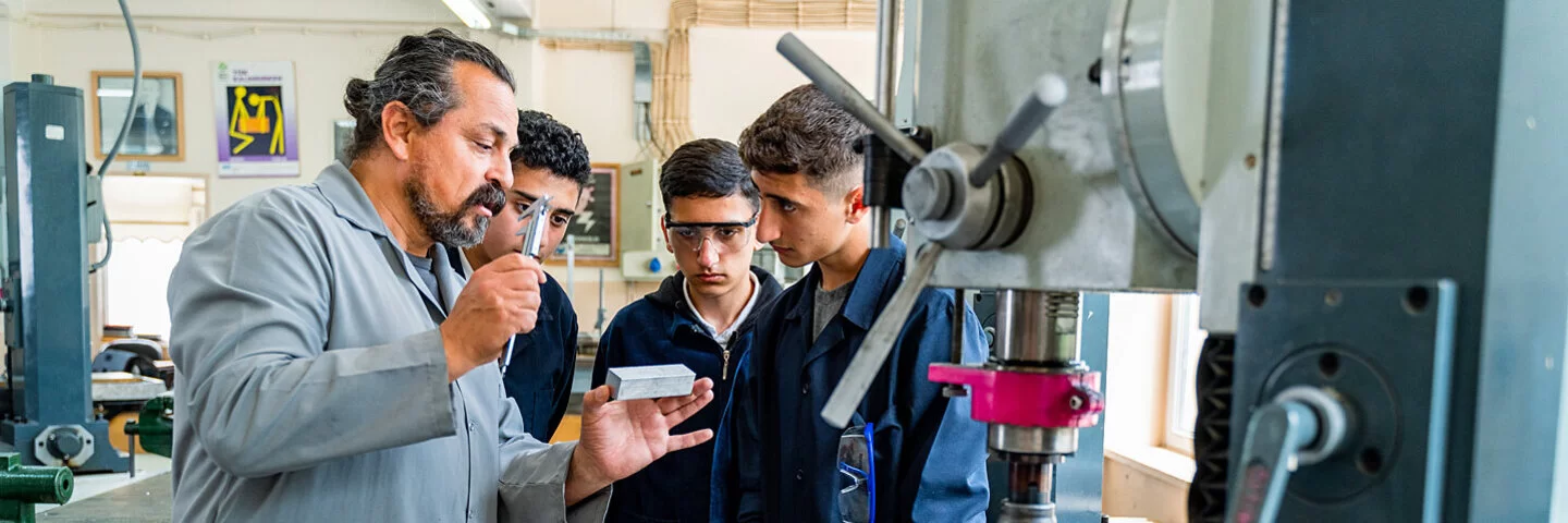 Ein Berufschullehrer erklärt in einer Feinmechanik-Werkstatt Auszubildenden ein Werkstück.