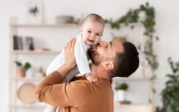 Ein junger Vater hebt in seinem Wohnzimmer sein Baby hoch und küsst es auf die Wange.