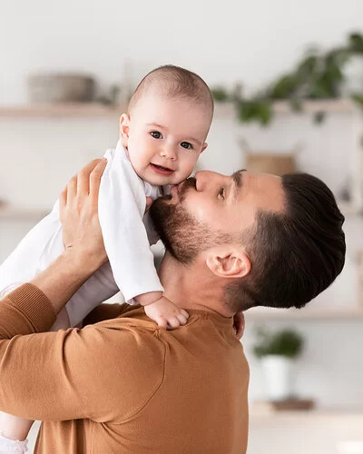 Ein junger Vater hebt in seinem Wohnzimmer sein Baby hoch und küsst es auf die Wange.