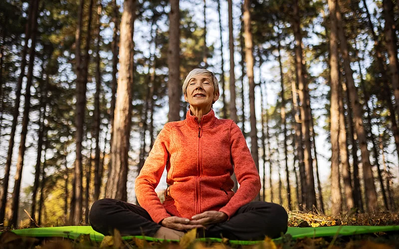 Selbsthilfe bei COPD: Eine Frau mittleren Alters sitzt auf ihrer Yoga-Matte mit geschlossenen Augen auf einer Lichtung im Wald und meditiert.