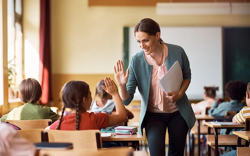 Eine Lehrerin gibt einem sitzenden Schulkind im Klassenzimmer ein High Five, während die anderen Kinder arbeiten.