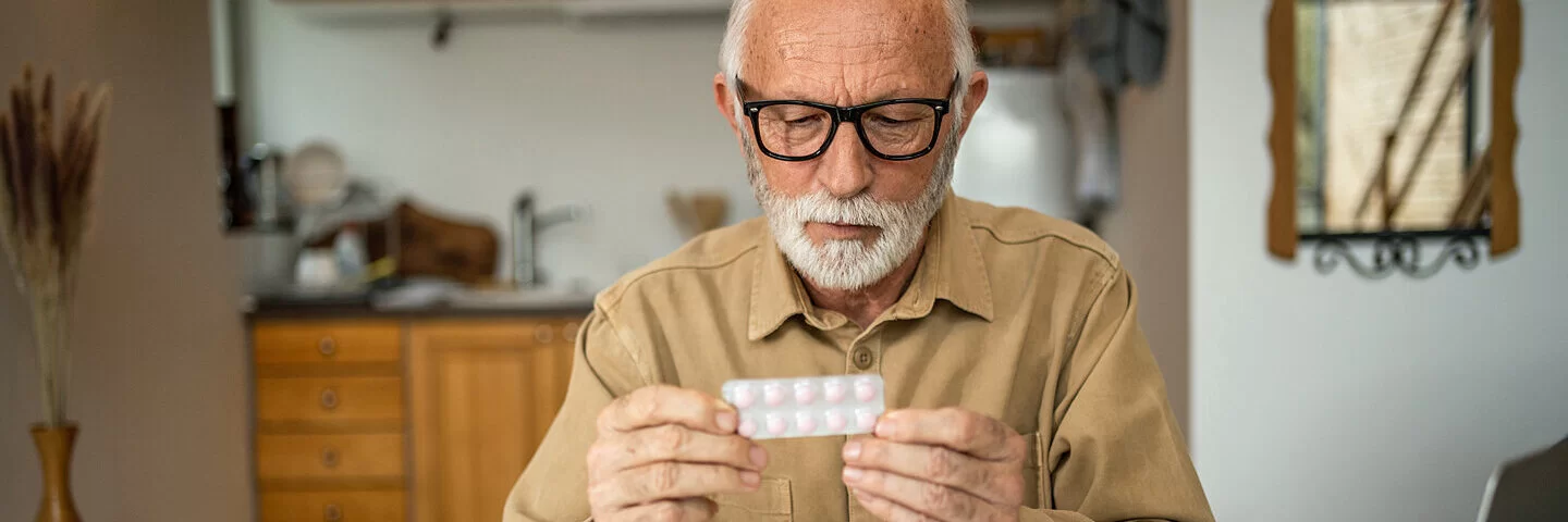 Ein alter Mann sitzt an seinem Küchentisch und schaut auf ein Tablettenblister in seinen Händen.