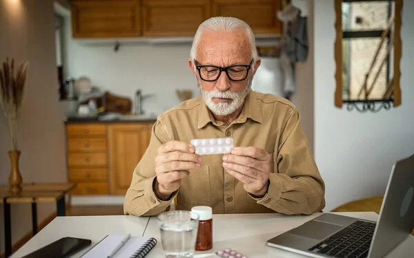Ein alter Mann sitzt an seinem Küchentisch und schaut auf ein Tablettenblister in seinen Händen.