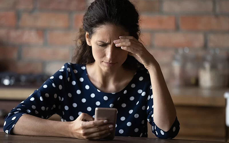 Eine Frau mit braunen Locken sitzt am Tisch. Sie fasst sich mit der linken Hand an die Stirn und schaut besorgt auf ihr Mobiltelefon.