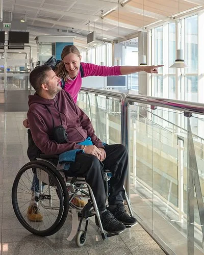 Ein Mann im Rollstuhl blickt am Flughafen aus dem Fenster. Seine Begleiterin streckt ihren linken Arm nach vorne, um ihm etwas zu zeigen.