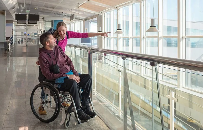 Ein Mann im Rollstuhl blickt am Flughafen aus dem Fenster. Seine Begleiterin streckt ihren linken Arm nach vorne, um ihm etwas zu zeigen.