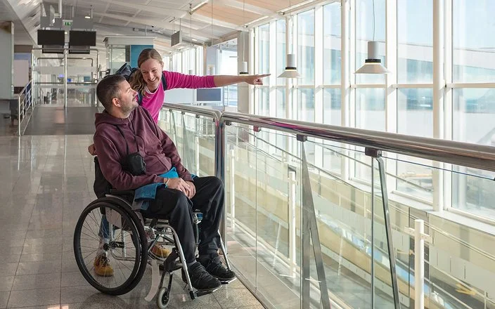 Ein Mann im Rollstuhl blickt am Flughafen aus dem Fenster. Seine Begleiterin streckt ihren linken Arm nach vorne, um ihm etwas zu zeigen.