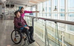 Ein Mann im Rollstuhl blickt am Flughafen aus dem Fenster. Seine Begleiterin streckt ihren linken Arm nach vorne, um ihm etwas zu zeigen.