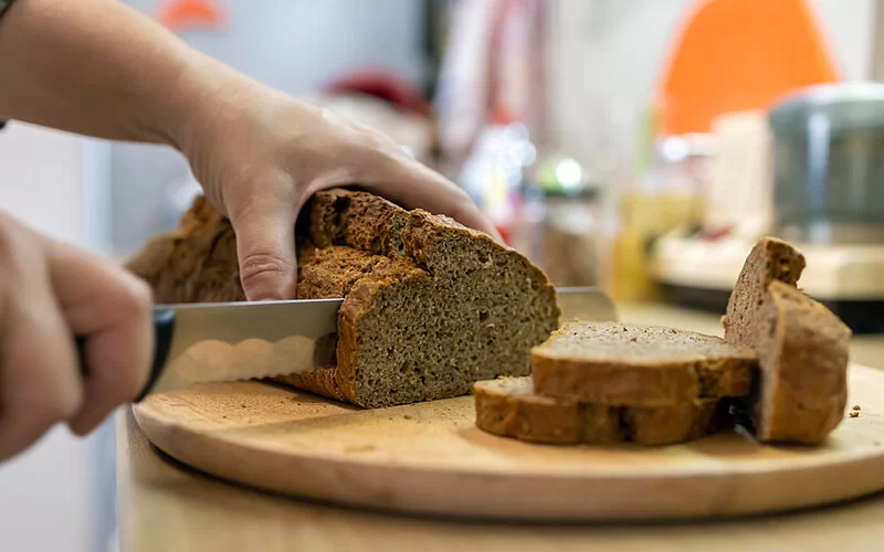 Eine Person schneidet mit einem Brotmesser ein frisches Vollkornbrot in Scheiben, Vollkorn ist ein guter Vitamin B1-Lieferant.