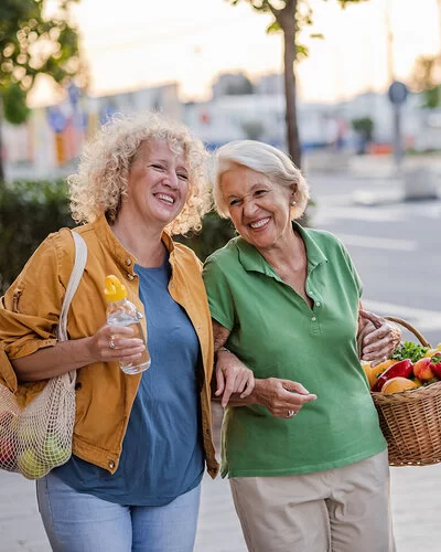 Zwei ältere blonde Frauen laufen untergehakt mit jeweils einem Einkaufskorb bzw. Einkaufsnetz voller Gemüse und Obst eine Straße entlang.