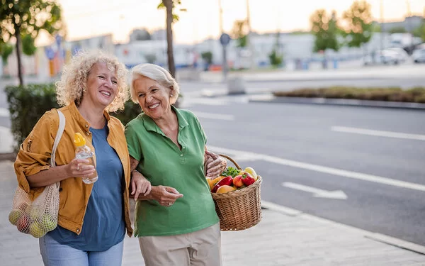 Zwei ältere blonde Frauen laufen untergehakt mit jeweils einem Einkaufskorb bzw. Einkaufsnetz voller Gemüse und Obst eine Straße entlang.