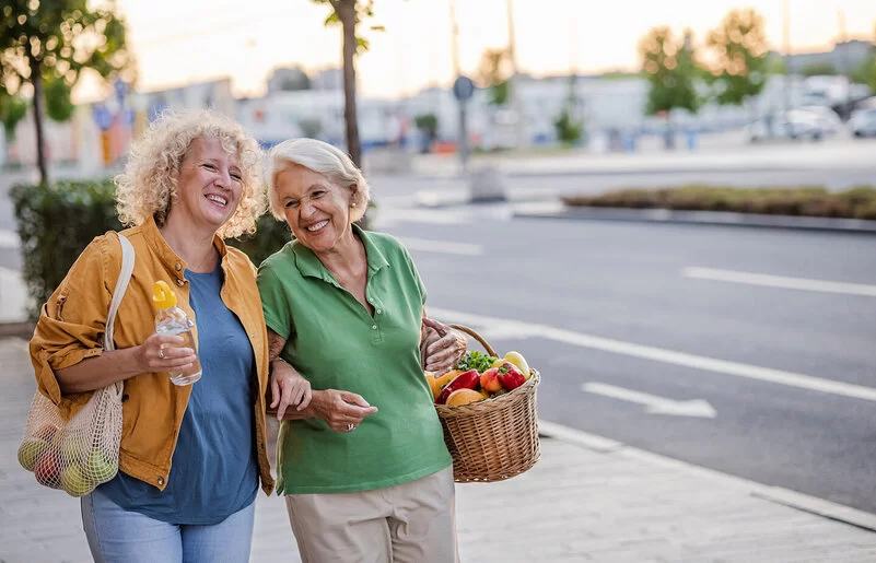 Zwei ältere blonde Frauen laufen untergehakt mit jeweils einem Einkaufskorb bzw. Einkaufsnetz voller Gemüse und Obst eine Straße entlang.