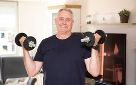 Auf dem Bild ist ein mittelalter Mann zu sehen, der in beiden Händen eine Hantel hält. 