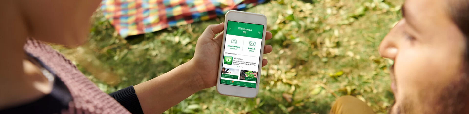 Eine Frau und ein Mann sitzen vor einer Picknickdecke und betrachten ein Smartphone mit einer darauf geöffneten AOK-App.