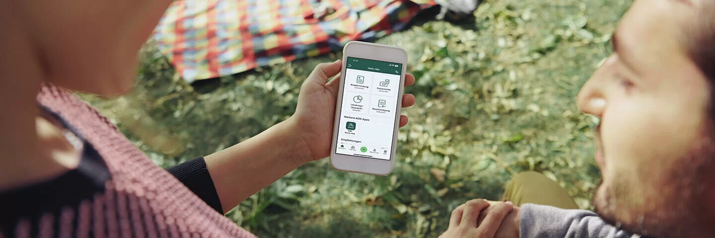 Eine Frau und ein Mann sitzen vor einer Picknickdecke und betrachten ein Smartphone mit einer darauf geöffneten AOK-App.