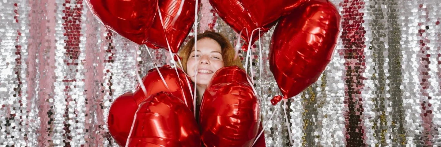 Glückliche Frau hält mehrere herzförmige Luftballons vor einem Paillettenvorhang.