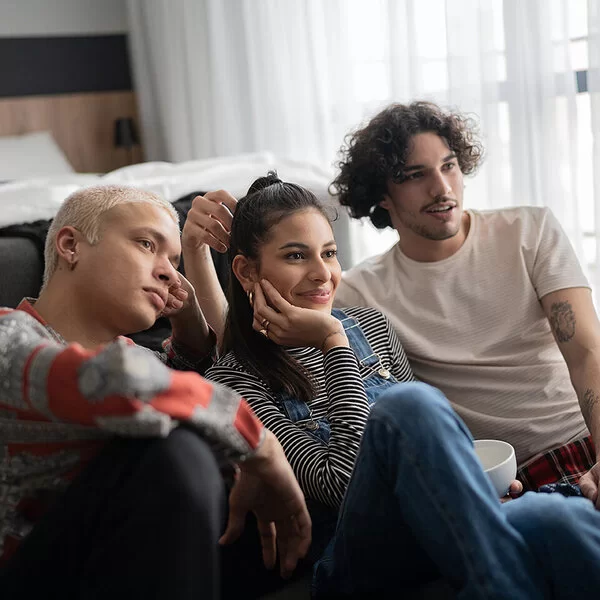 Zwei männliche und eine weibliche Person sitzen zusammen auf dem Fußboden und sehen fern.