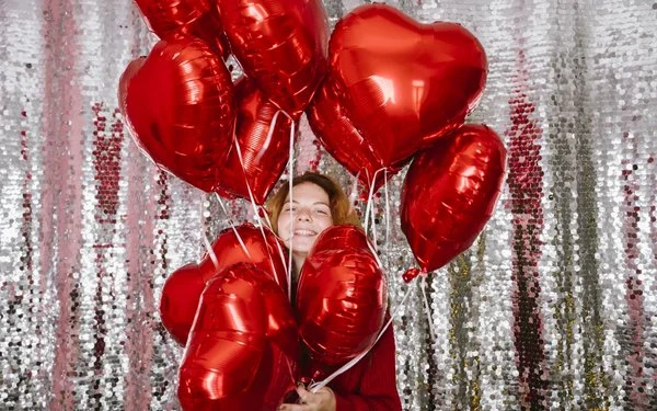 Glückliche Frau hält mehrere herzförmige Luftballons vor einem Paillettenvorhang.