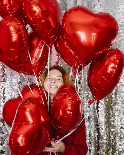 Glückliche Frau hält mehrere herzförmige Luftballons vor einem Paillettenvorhang.