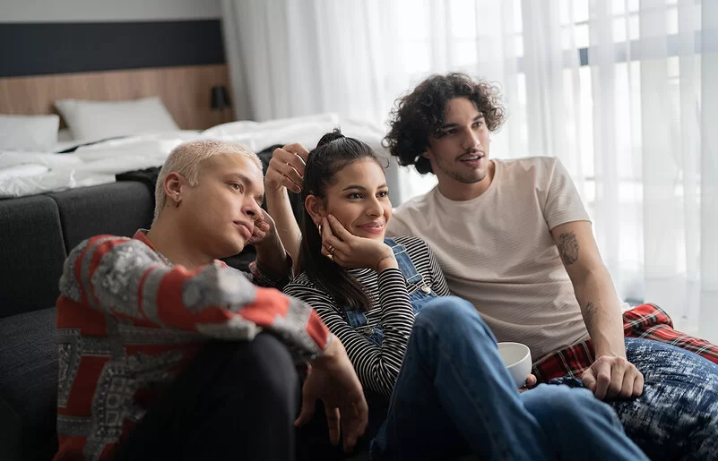 Zwei männliche und eine weibliche Person sitzen zusammen auf dem Fußboden und sehen fern.