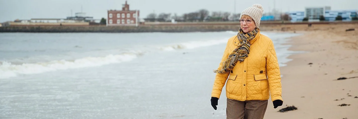 Ältere Frau mit dicker Jacke, Mütze und Schal geht am Meer spazieren, im Hintergrund sind Häuser zu sehen.