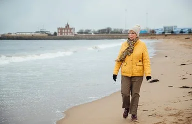 Ältere Frau mit dicker Jacke, Mütze und Schal geht am Meer spazieren, im Hintergrund sind Häuser zu sehen.