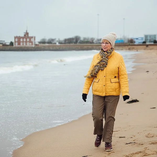 Ältere Frau mit dicker Jacke, Mütze und Schal geht am Meer spazieren, im Hintergrund sind Häuser zu sehen.