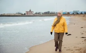 Ältere Frau mit dicker Jacke, Mütze und Schal geht am Meer spazieren, im Hintergrund sind Häuser zu sehen.