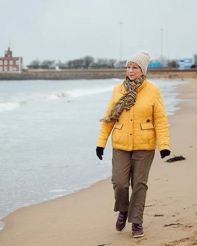 Ältere Frau mit dicker Jacke, Mütze und Schal geht am Meer spazieren, im Hintergrund sind Häuser zu sehen.