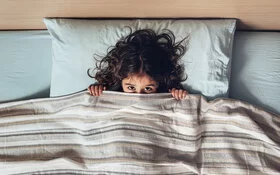 Ein kleines Mädchen liegt ängstlich im Bett und zieht die Bettdecke über das Gesicht, sodass nur die Augen hervorgucken.
