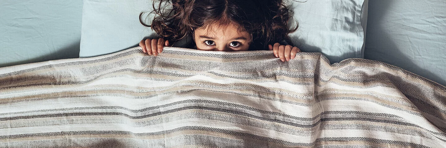 Ein kleines Mädchen liegt ängstlich im Bett und zieht die Bettdecke über das Gesicht, sodass nur die Augen hervorgucken.