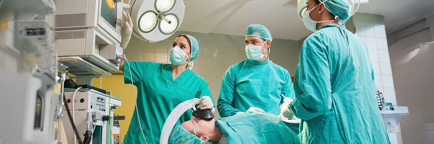 Ein Operationssaal. Zwei Ärztinnen und ein Arzt in grüner OP-Kleidung betrachten ein technisches Gerät. Eine Patientin auf dem Operationstisch wird über eine Beatmungsmaske beatmet.