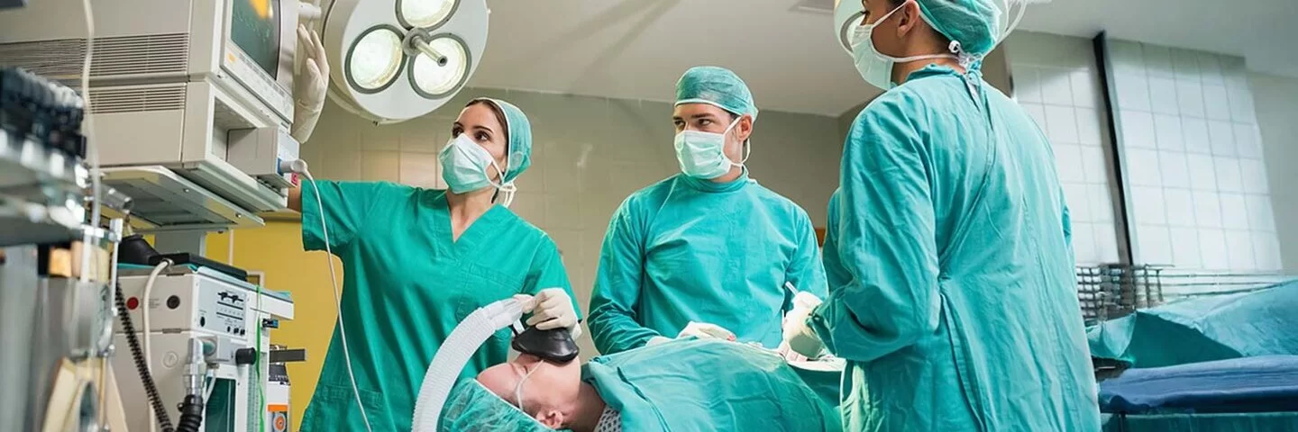 Ein Operationssaal. Zwei Ärztinnen und ein Arzt in grüner OP-Kleidung betrachten ein technisches Gerät. Eine Patientin auf dem Operationstisch wird über eine Beatmungsmaske beatmet.