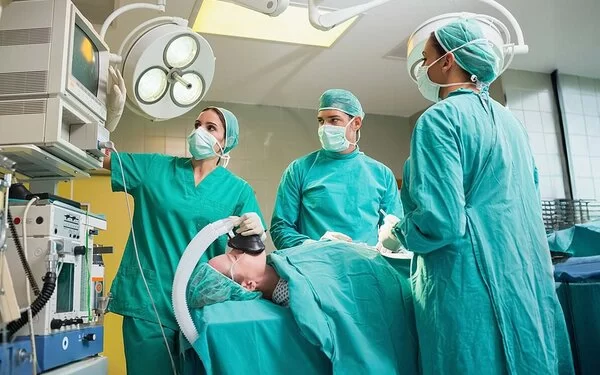 Ein Operationssaal. Zwei Ärztinnen und ein Arzt in grüner OP-Kleidung betrachten ein technisches Gerät. Eine Patientin auf dem Operationstisch wird über eine Beatmungsmaske beatmet.